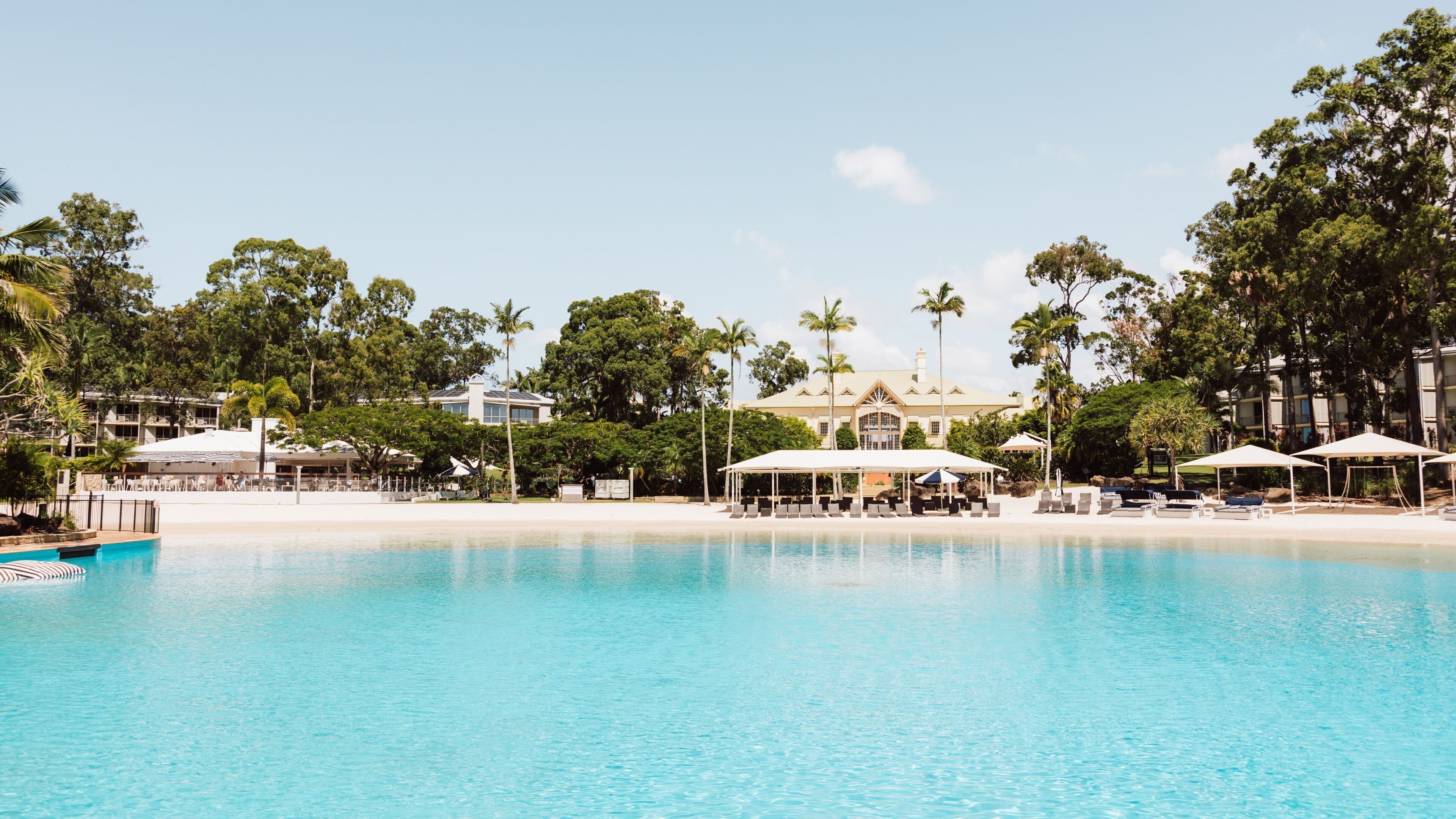 A pool with lounge chairs and umbrellas, surrounded by palm trees and a large building with a white roof.
