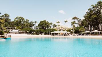 A pool with lounge chairs and umbrellas, surrounded by palm trees and a large building with a white roof.