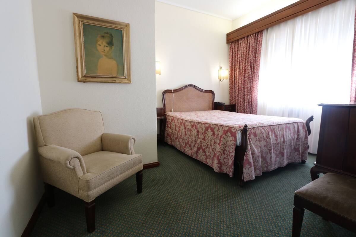 A hotel room with a single bed, a chair, a desk, and a painting on the wall.