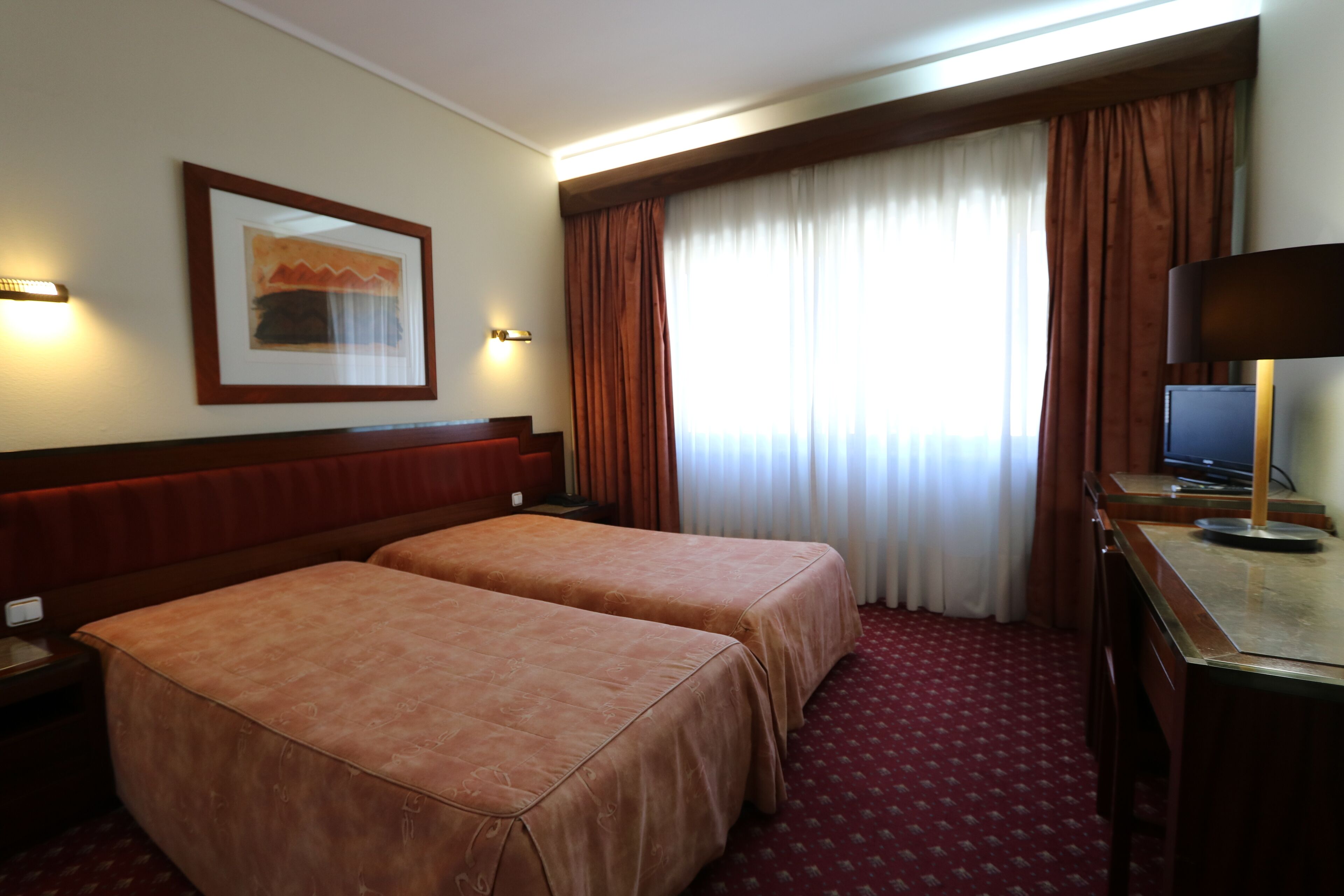 A hotel room with two beds, a desk with a computer, a framed picture on the wall, and a window with curtains.