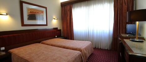 A hotel room with two beds, a desk with a computer, a framed picture on the wall, and a window with curtains.