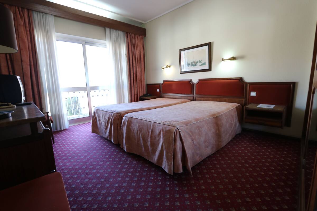 A hotel room with two beds, a desk, a chair, a window with curtains, and a framed picture on the wall.