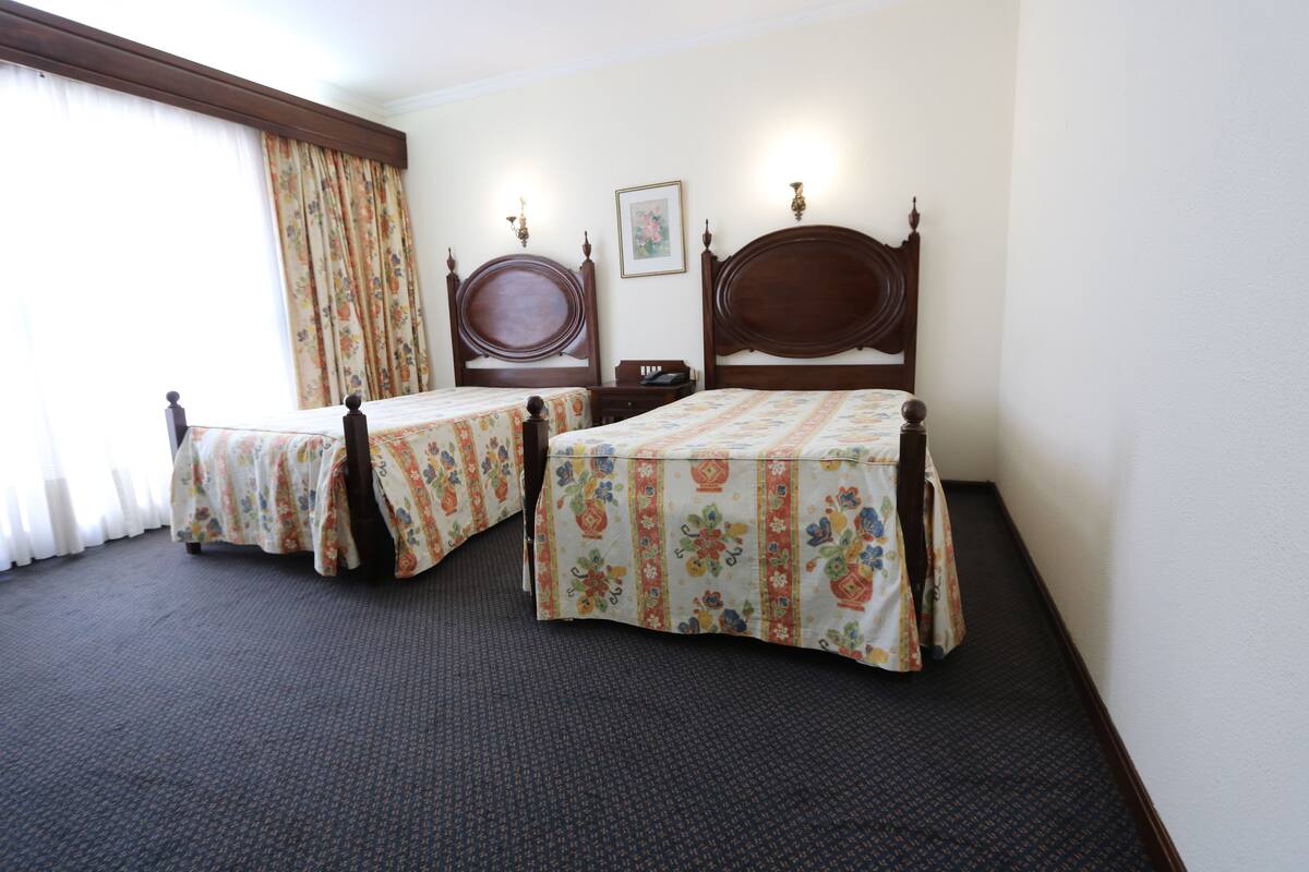 A hotel room with two single beds, a wooden headboard, a small bedside table, a nightstand, and a window with curtains.