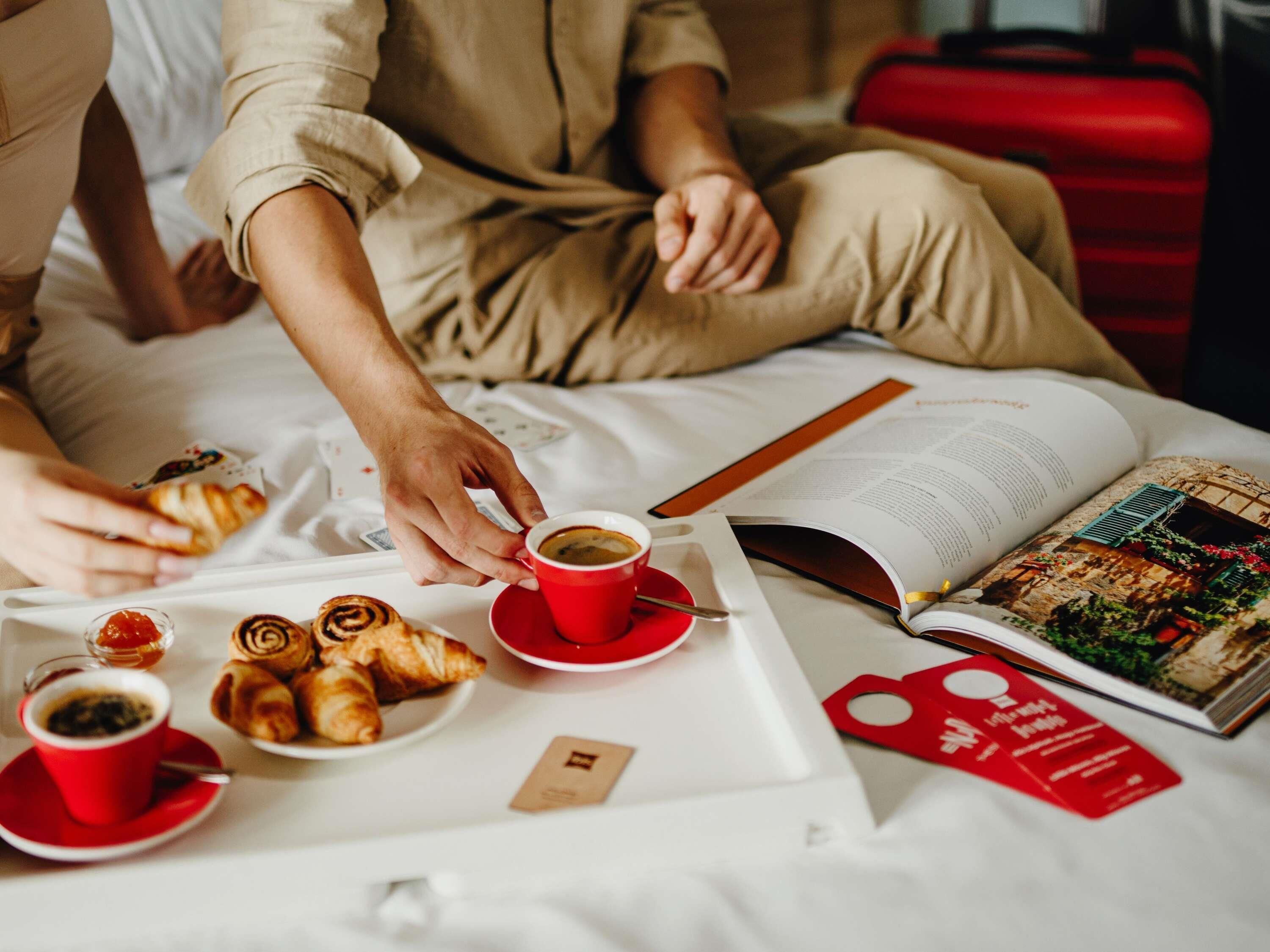 A bed with a tray of pastries and coffee, a book, and a red suitcase.