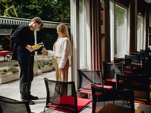 A man pouring a drink into a glass, a woman holding a glass, and outdoor seating with chairs and tables. - ibis Budapest Citysouth (Budapest)