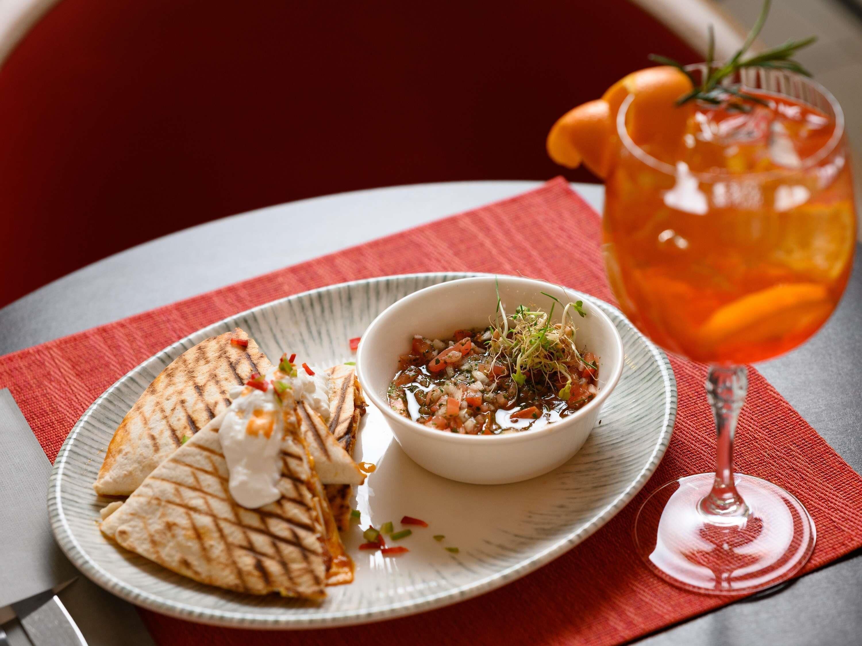 A plate with grilled quesadillas and a bowl of salsa, a glass of orange juice, and a red napkin on a table.