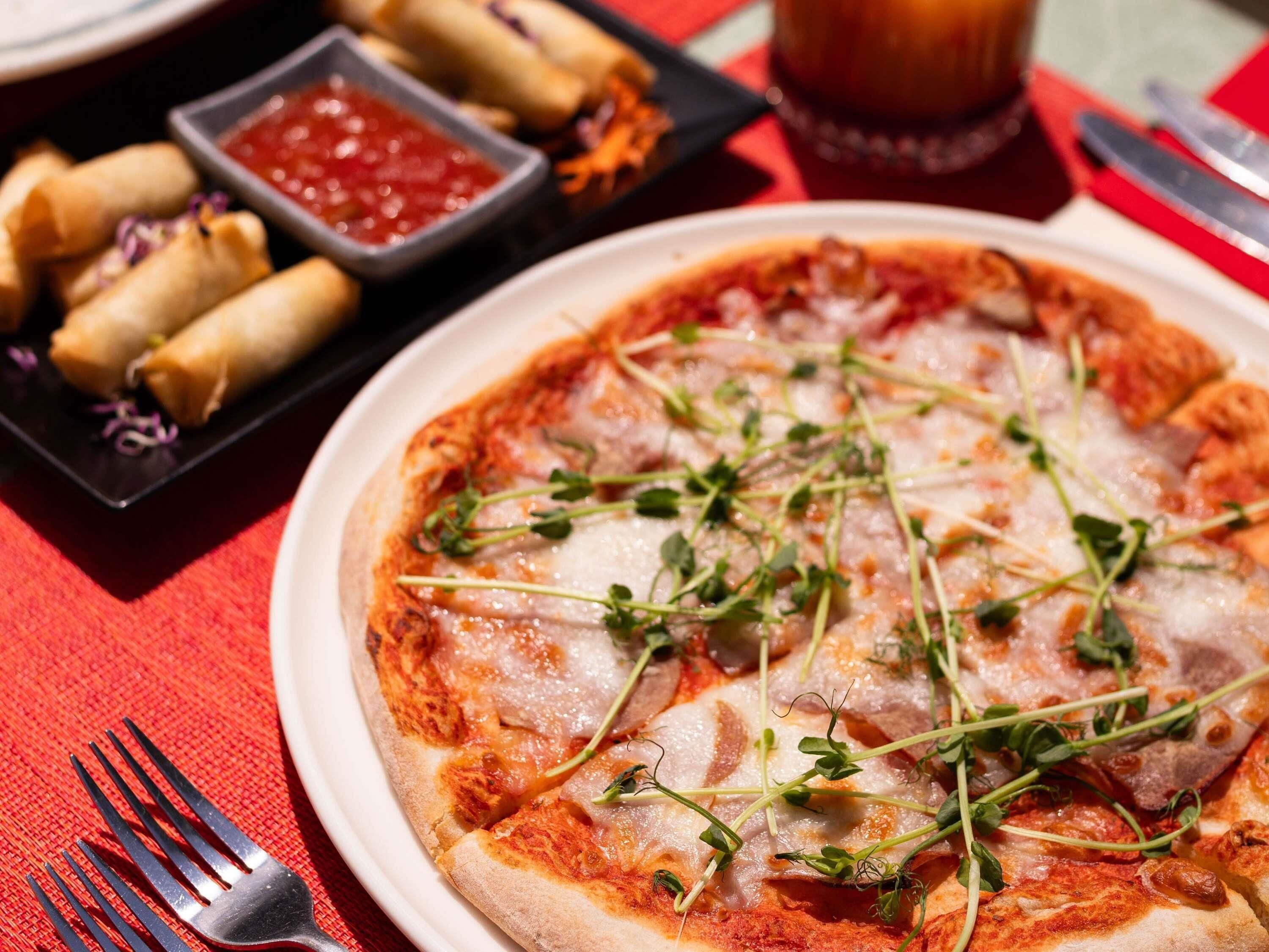 A pizza with cheese and greens on a plate, a fork, and a small bowl of sauce on a table.