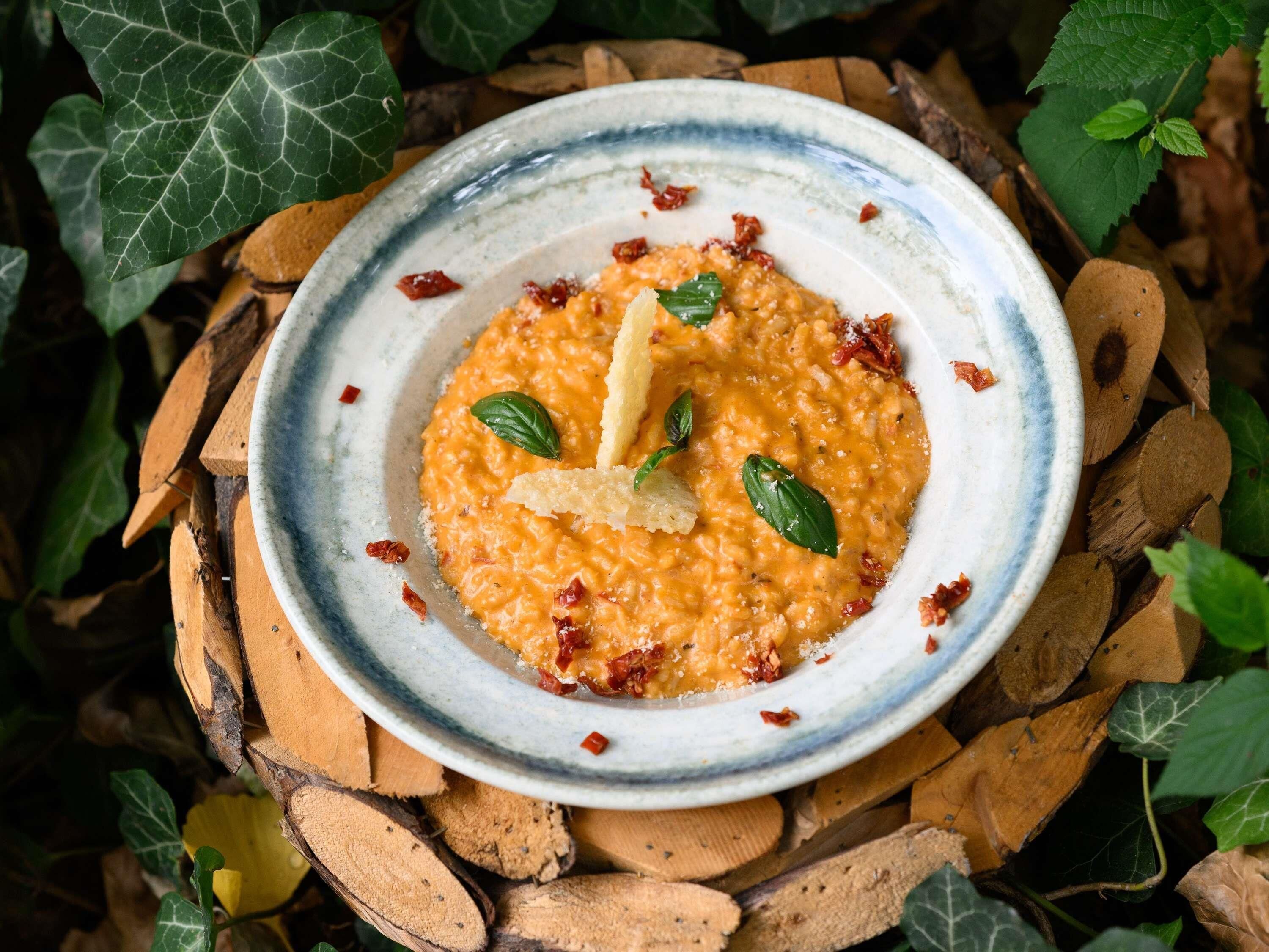 A bowl of orange-colored food with basil leaves and grated cheese on a rustic wooden surface.