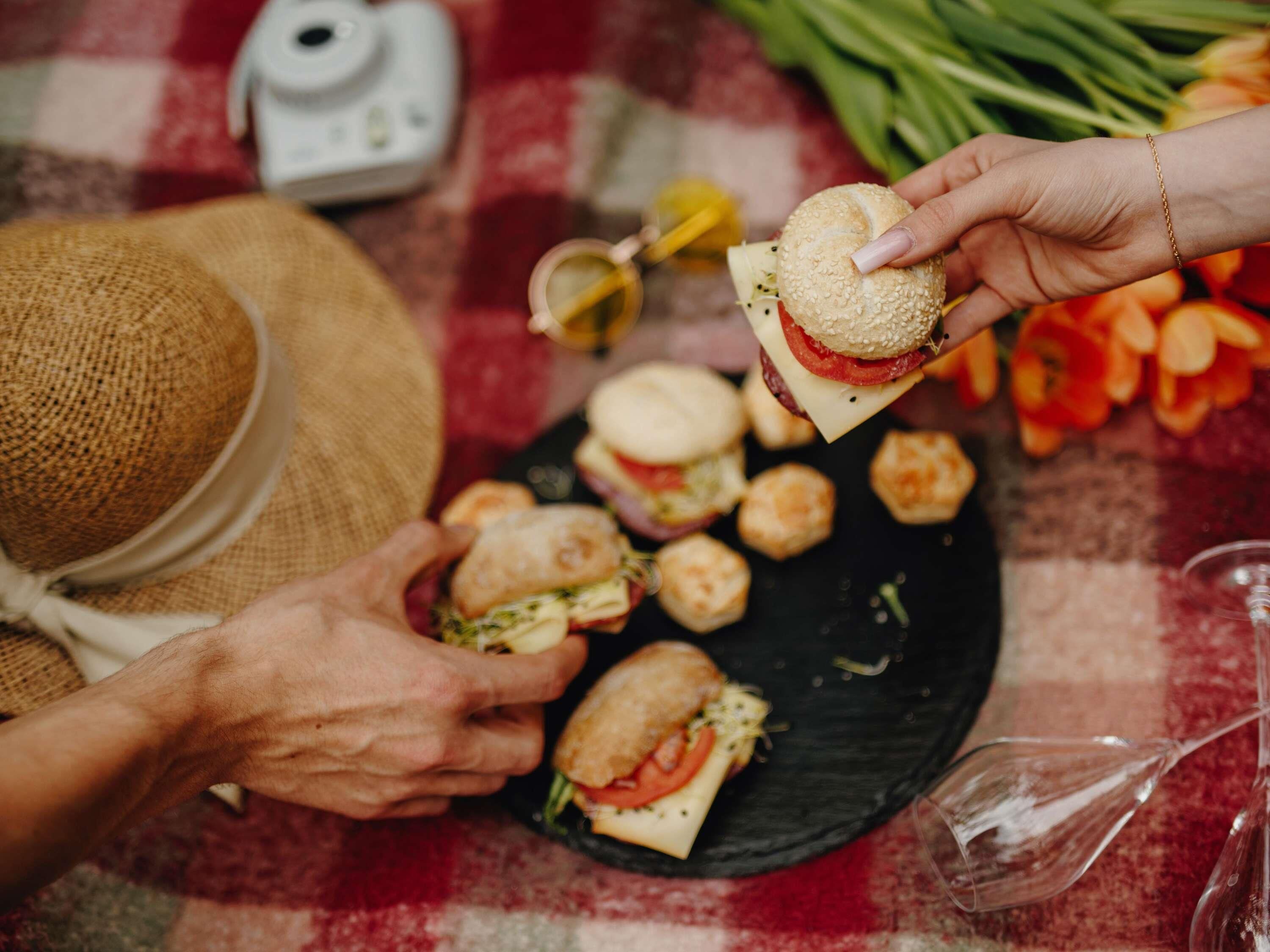 A person holding a sandwich, another person receiving it, and a plate with additional sandwiches.