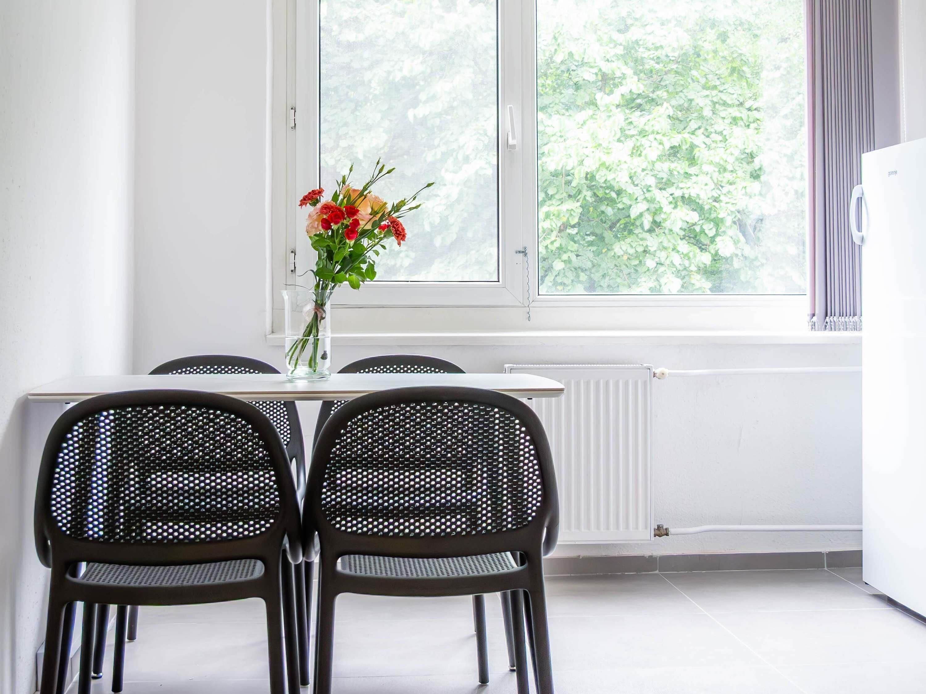 A room with a dining table, four chairs, a vase with flowers, a window with a view of trees, and a radiator.