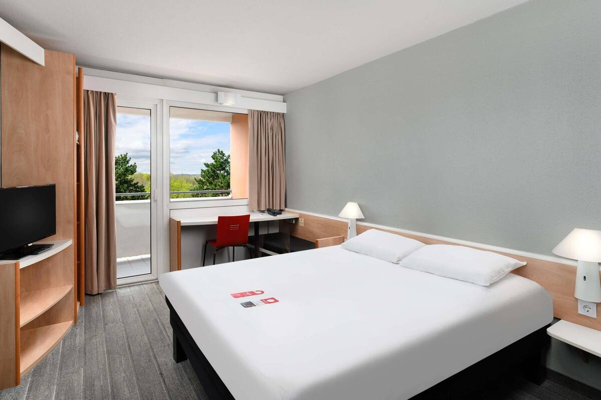 Habitación de hotel con cama, escritorio, silla, televisión y ventana con cortinas.