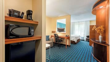 Une chambre d’hôtel équipée d’un four à micro-ondes, d’une machine à café et d’un petit réfrigérateur posé sur une étagère. On y trouve un bureau avec une chaise, un lit et une fenêtre avec des rideaux. La chambre est recouverte d’un tapis à motifs bleus.