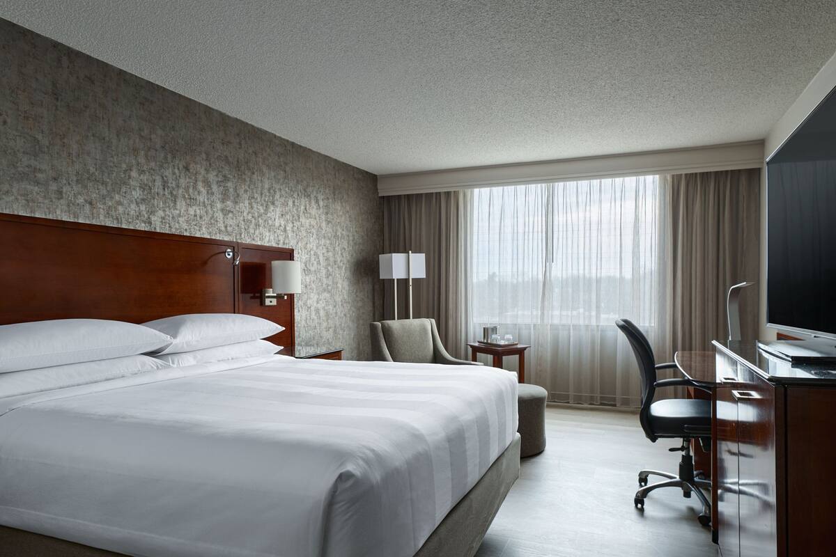 A hotel room with a large bed, a desk with a chair, a lamp, and a TV.