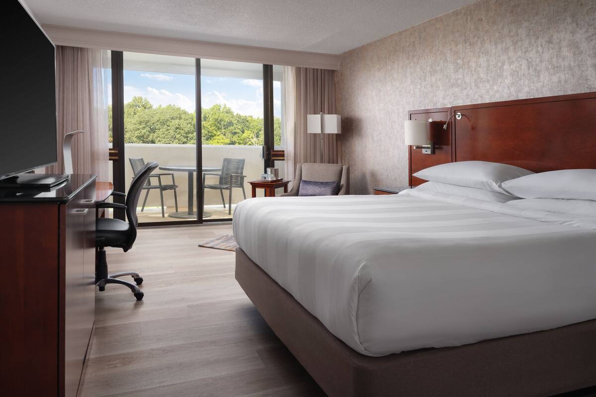 A hotel room with a large bed, a desk with a chair, a TV, and a balcony with a view of trees.