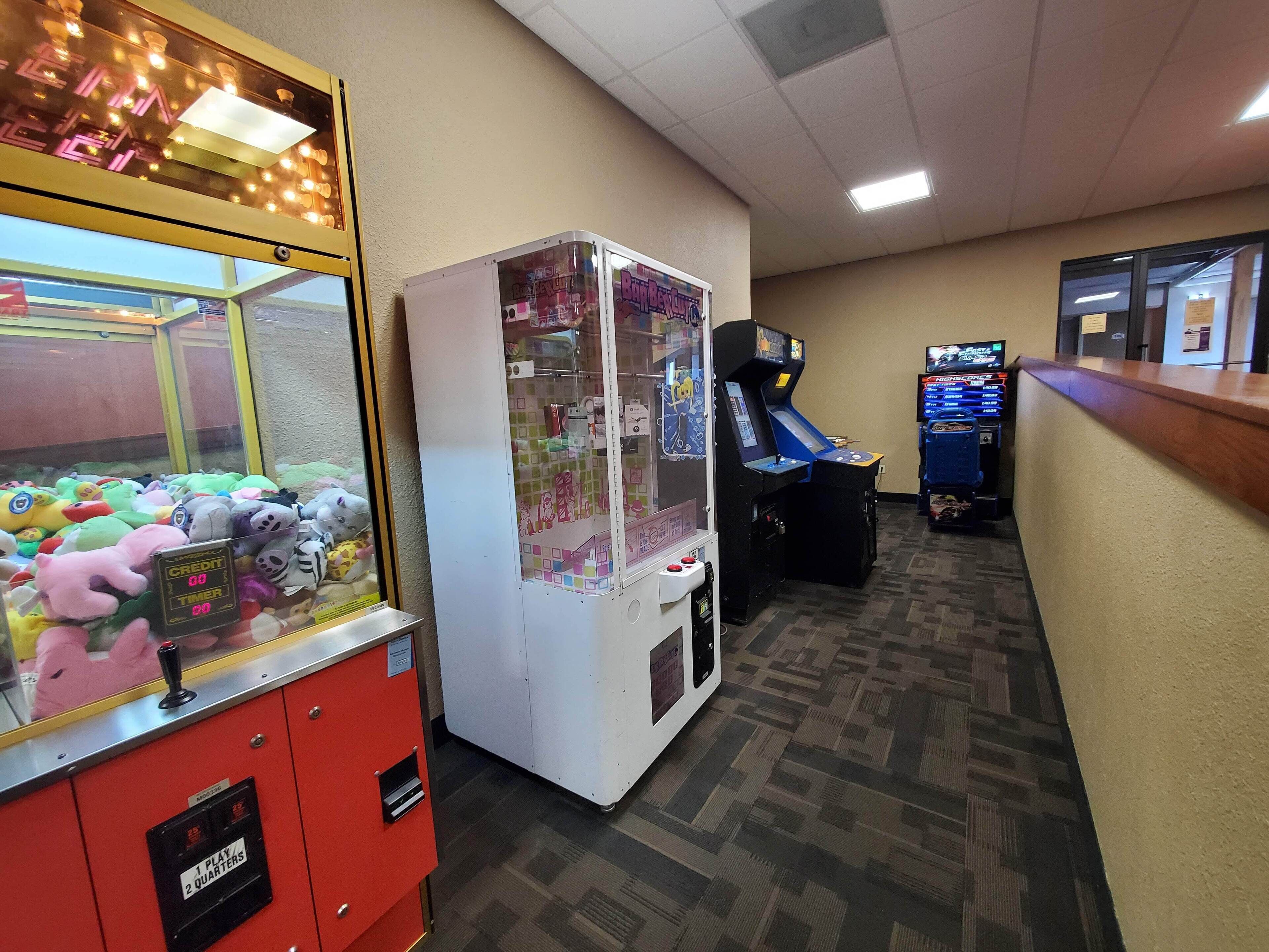 A hotel lobby with a claw machine filled with plush toys, a racing game machine, and a counter with a cash register.