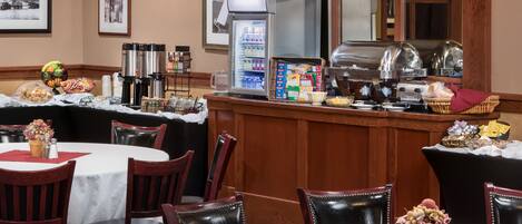 A hotel dining area with round tables, chairs, a buffet counter, and a refrigerator stocked with beverages.