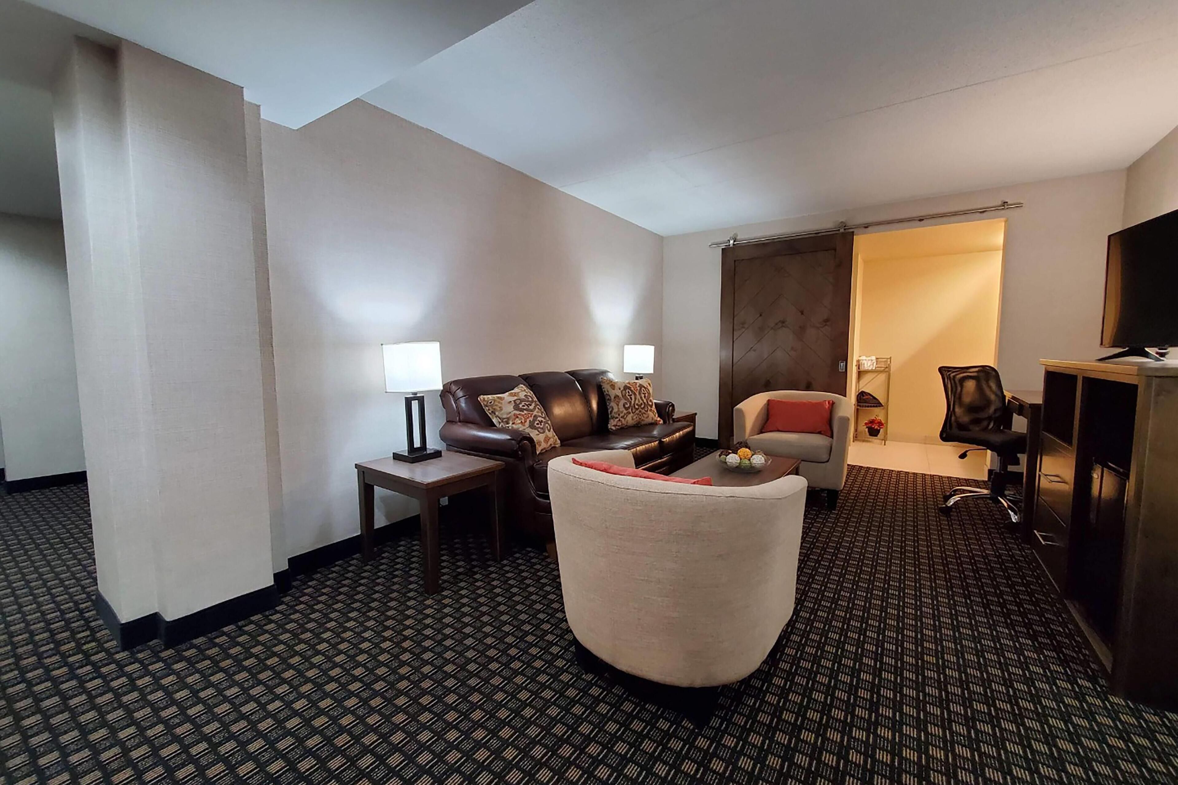 A hotel room with a brown leather sofa, a white armchair, a small table with a lamp, a TV, and a desk with a chair.