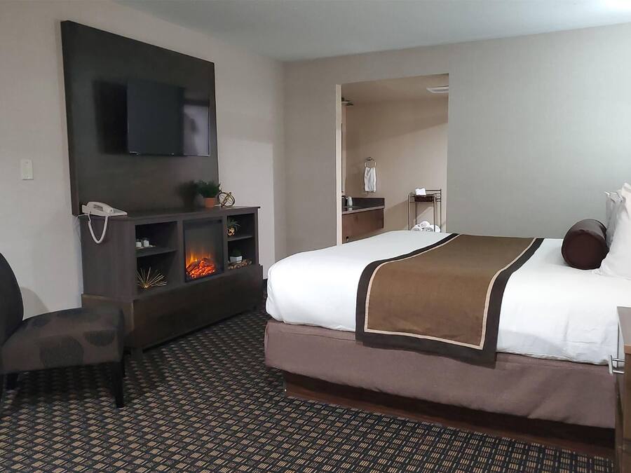 A hotel room with a bed, a TV mounted on the wall, a small seating area with a chair, a fireplace, and a bathroom visible through an open door.