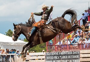 Evento de rodeo con un jinete sobre un caballo salvaje, espectadores de fondo y un cartel que indica el nombre y los reconocimientos del evento.