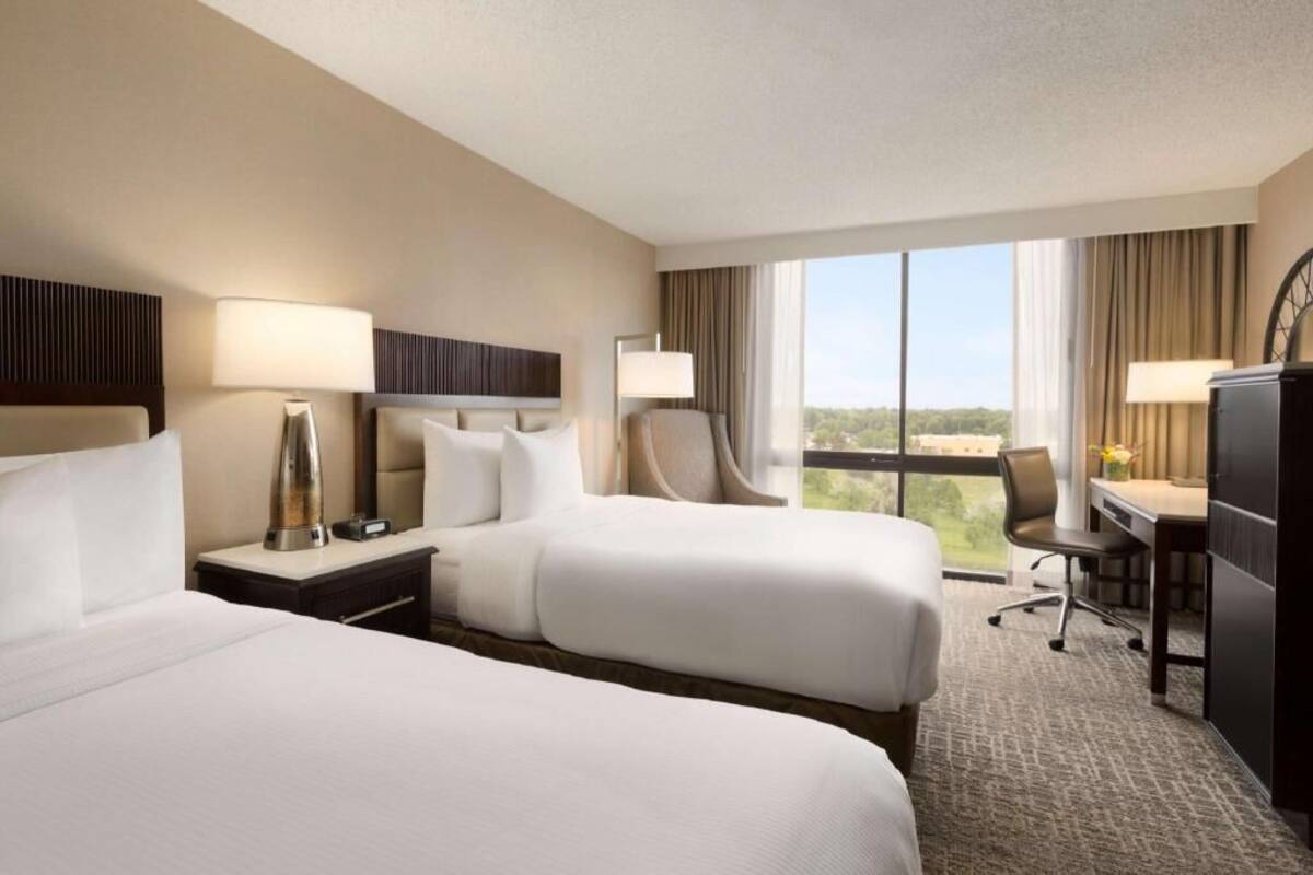 Quarto de hotel com duas camas, uma escrivaninha, uma cadeira e uma janela grande com vista.