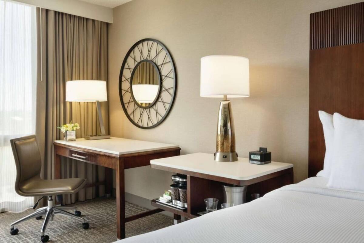 Quarto de hotel com cama, duas mesas de cabeceira, escrivaninha, cadeira, abajur, relógio e espelho.