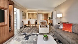 A modern living room with a sofa, armchair, and coffee table. There is a dining area with chairs and a table, and a kitchen in the background. - Hilton Fort Collins (Fort Collins)