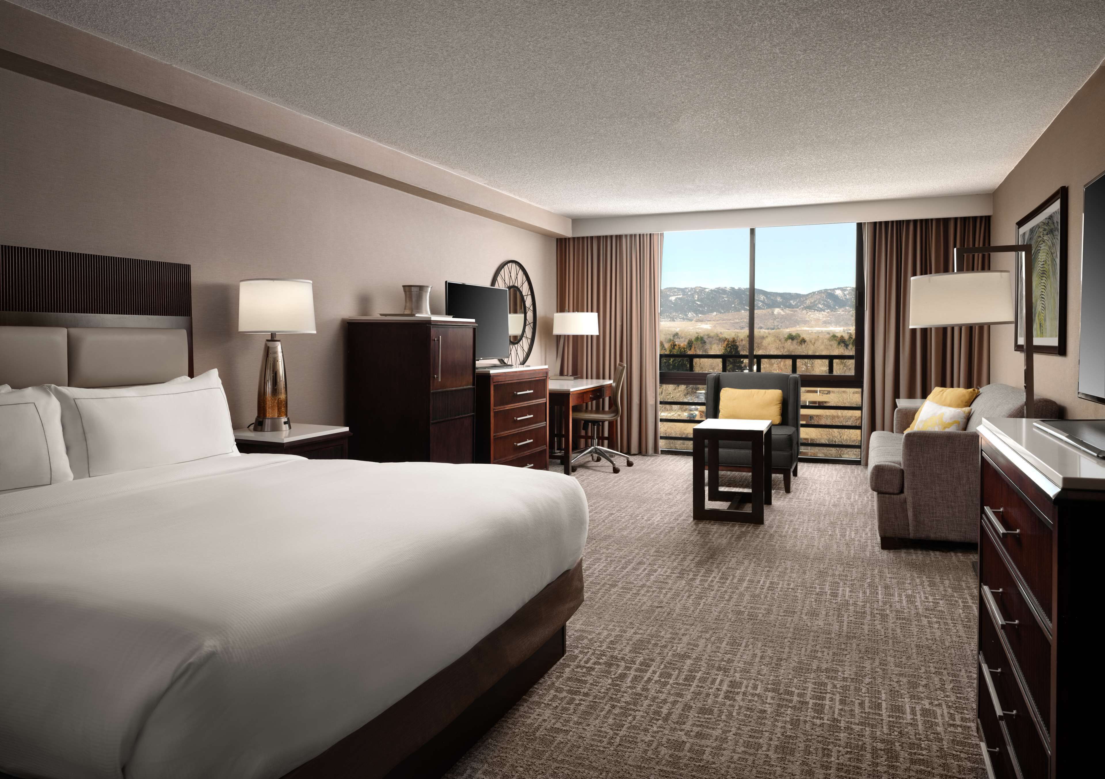 A hotel room with a large bed, a desk, a chair, a TV, and a view of the mountains.