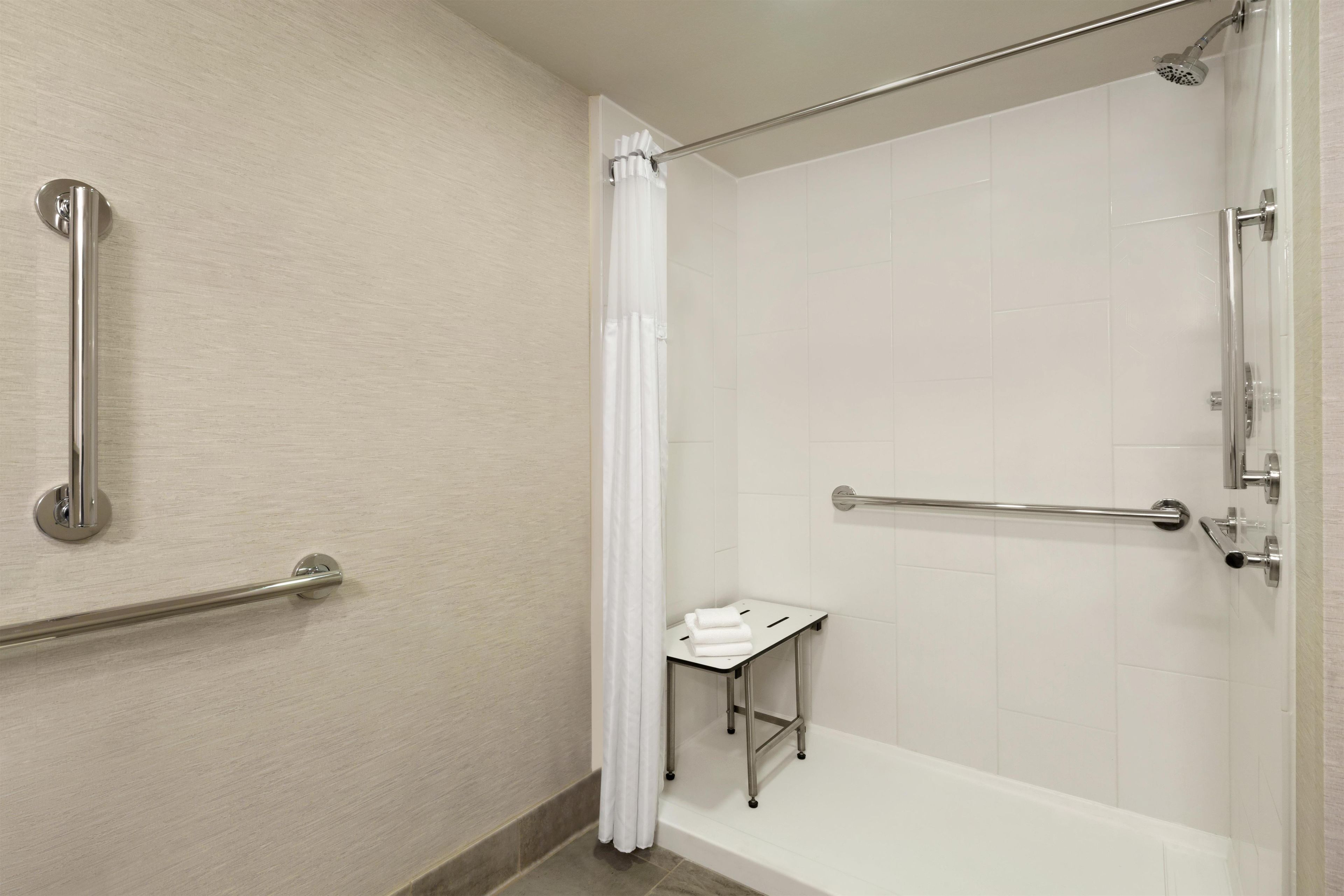 Une salle de bain avec une douche à l’italienne, des barres d’appui, un rideau de douche blanc et une petite table avec des serviettes.