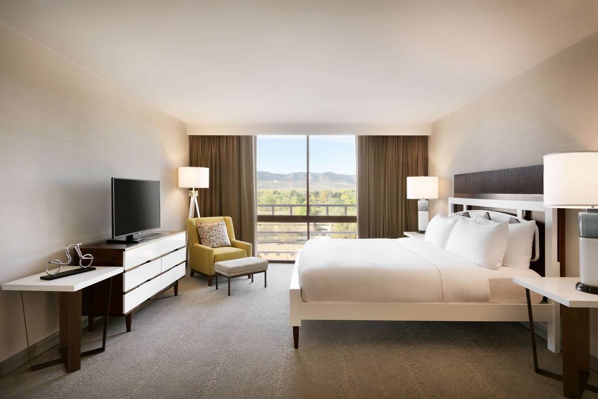 Quarto de hotel com cama, escrivaninha com televisão, cadeira e vista para as montanhas.