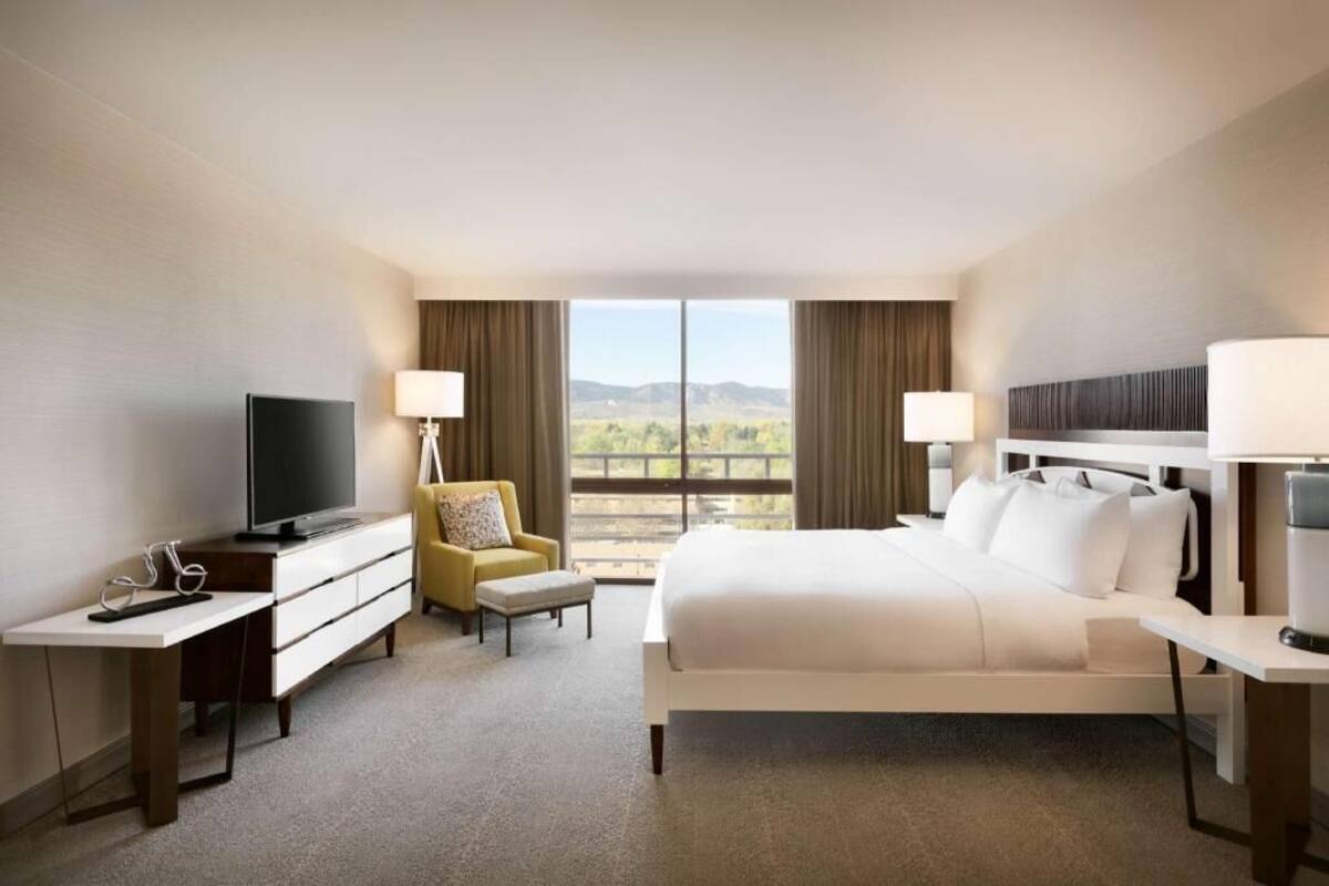 Quarto de hotel com cama, escrivaninha, cadeira, televisão e janela com vista.