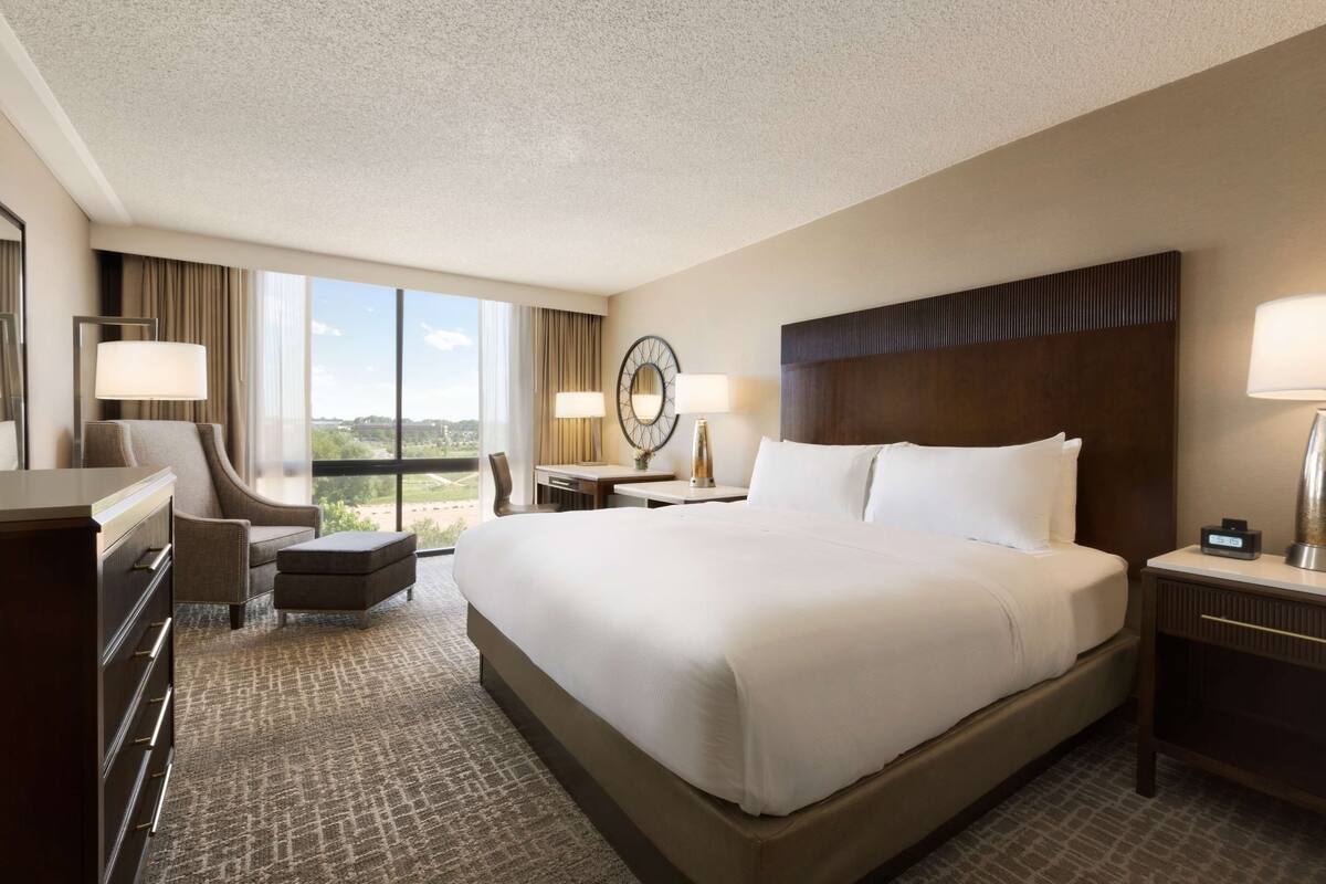 Quarto de hotel com uma cama grande, uma escrivaninha, uma cadeira, uma cômoda e vista para o exterior.