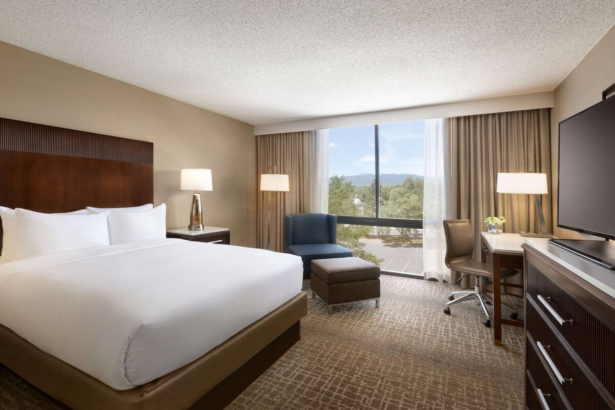 Quarto de hotel com uma cama grande, uma escrivaninha com cadeira, televisão e vista para o exterior através de amplas janelas.
