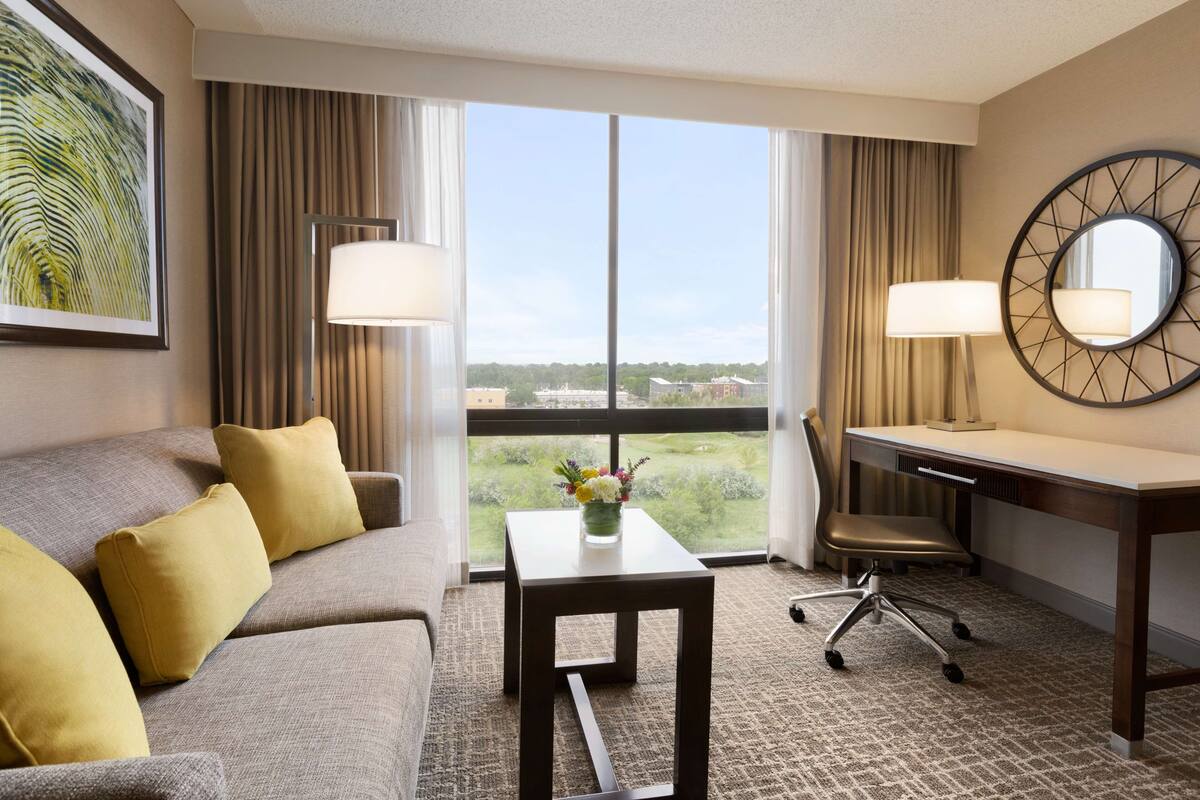 Quarto de hotel com sofá, escrivaninha, cadeira, mesinha de centro com um vaso e uma janela grande com vista para o exterior.