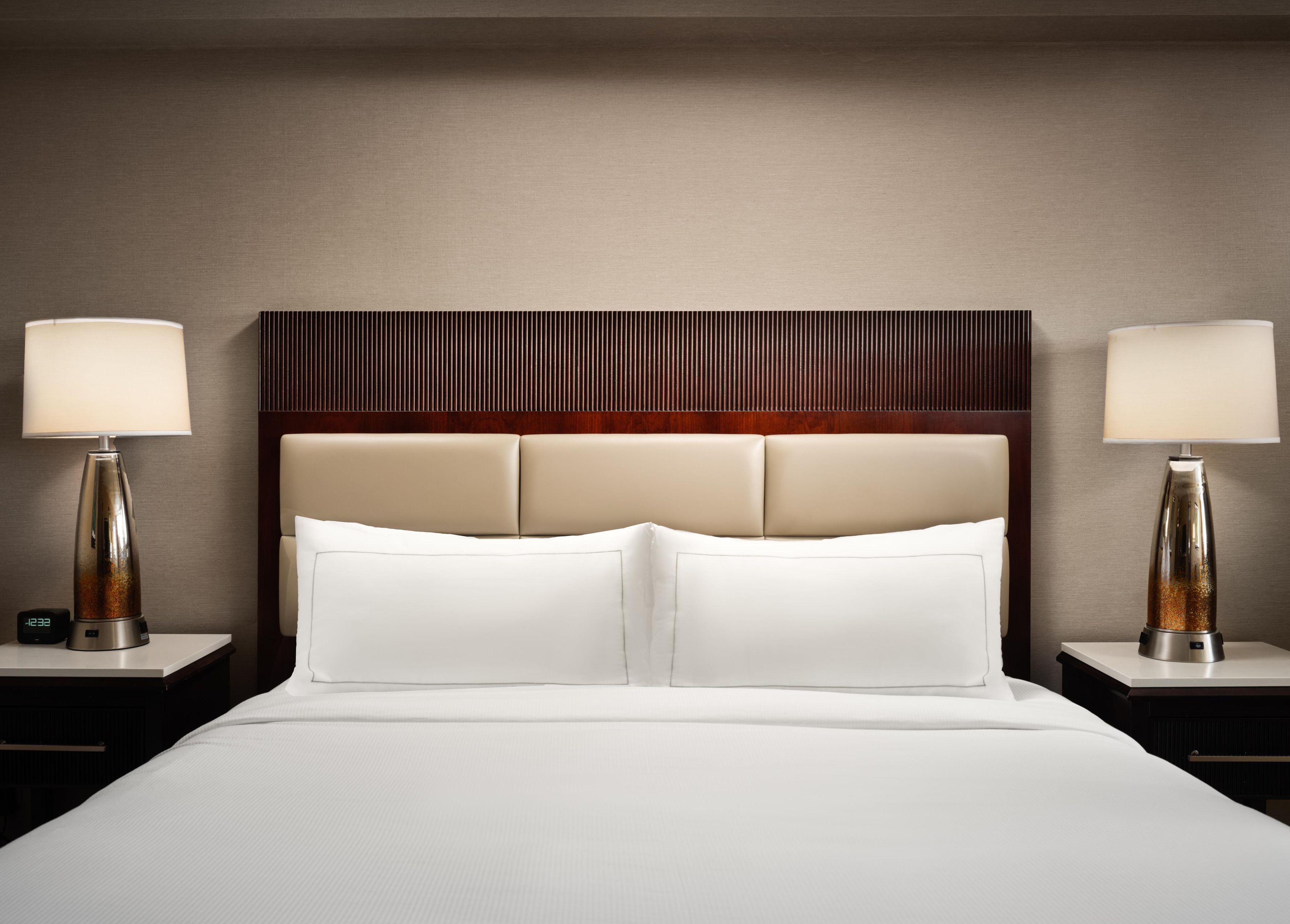 A hotel room with a bed, two bedside lamps, a digital clock, and a wooden headboard.