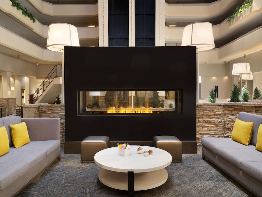 A spacious lobby with a central fireplace, two sofas with yellow cushions, a round coffee table, and a stone accent wall.