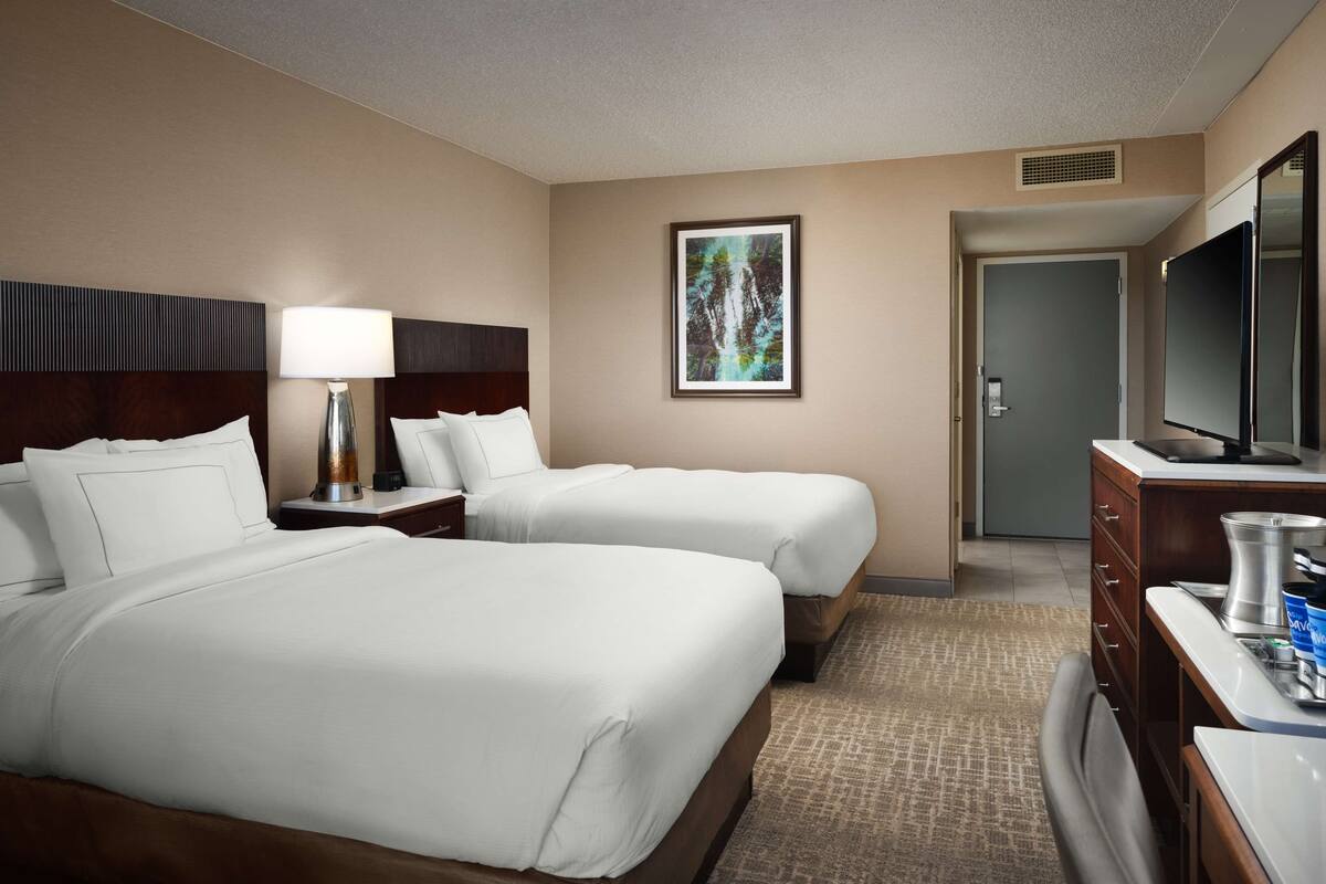 Quarto de hotel com duas camas, televisão, escrivaninha com luminária, um quadro na parede e porta de acesso a um banheiro.