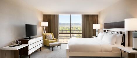 A hotel room with a bed, a desk, a television, a chair, and a view of the mountains.