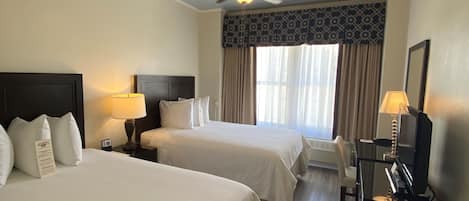 A hotel room with two beds, a ceiling fan, a desk with a lamp, a chair, and a television.