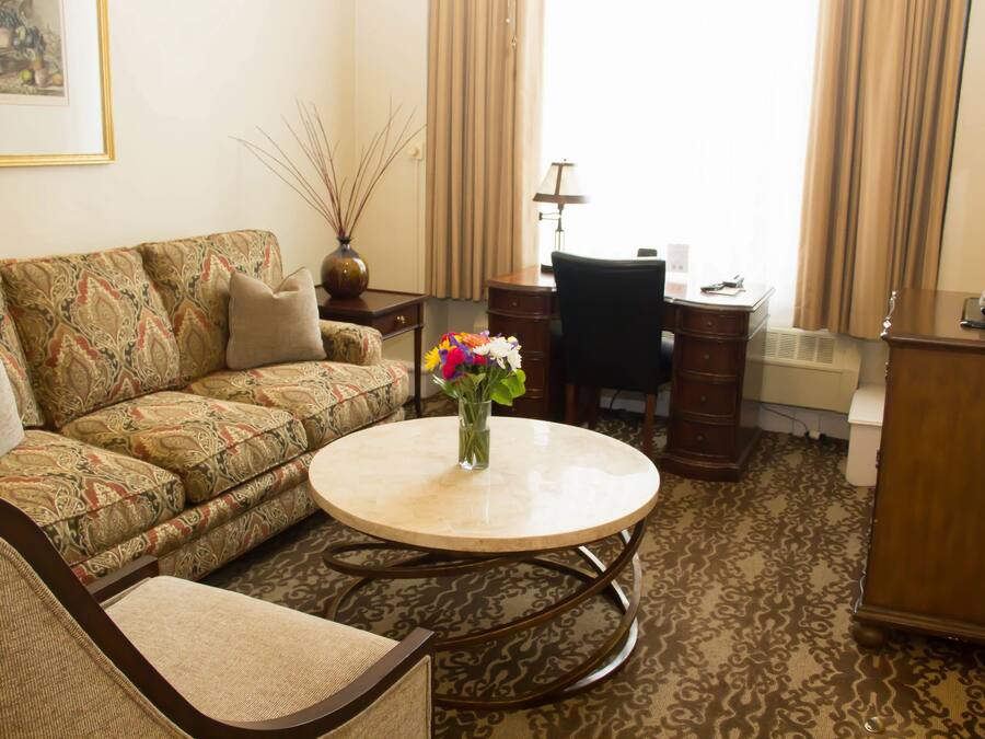 A living room with a patterned sofa, a round coffee table with a vase of flowers, a desk with a chair, and a television on a wooden cabinet.