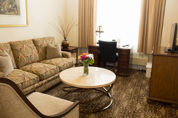 A living room with a patterned sofa, a round coffee table with a vase of flowers, a desk with a chair, and a television on a wooden cabinet. - Chancellor Hotel on Union Square (San Francisco)