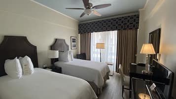Quarto de hotel com duas camas, um ventilador de teto, uma mesa com abajur e vista para o exterior através de grandes janelas.