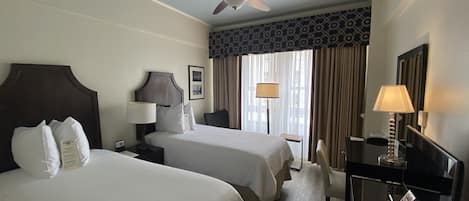 Habitación de hotel con dos camas, un ventilador de techo, un escritorio con lámpara y vistas al exterior a través de amplias ventanas.