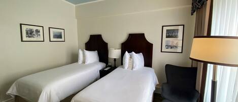 A hotel room with two single beds, a ceiling fan, a chair, and framed pictures on the wall.