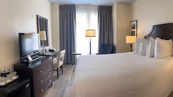 Habitación de hotel con una cama grande, un escritorio con silla, televisor, minibar y una lámpara en la mesita de noche.