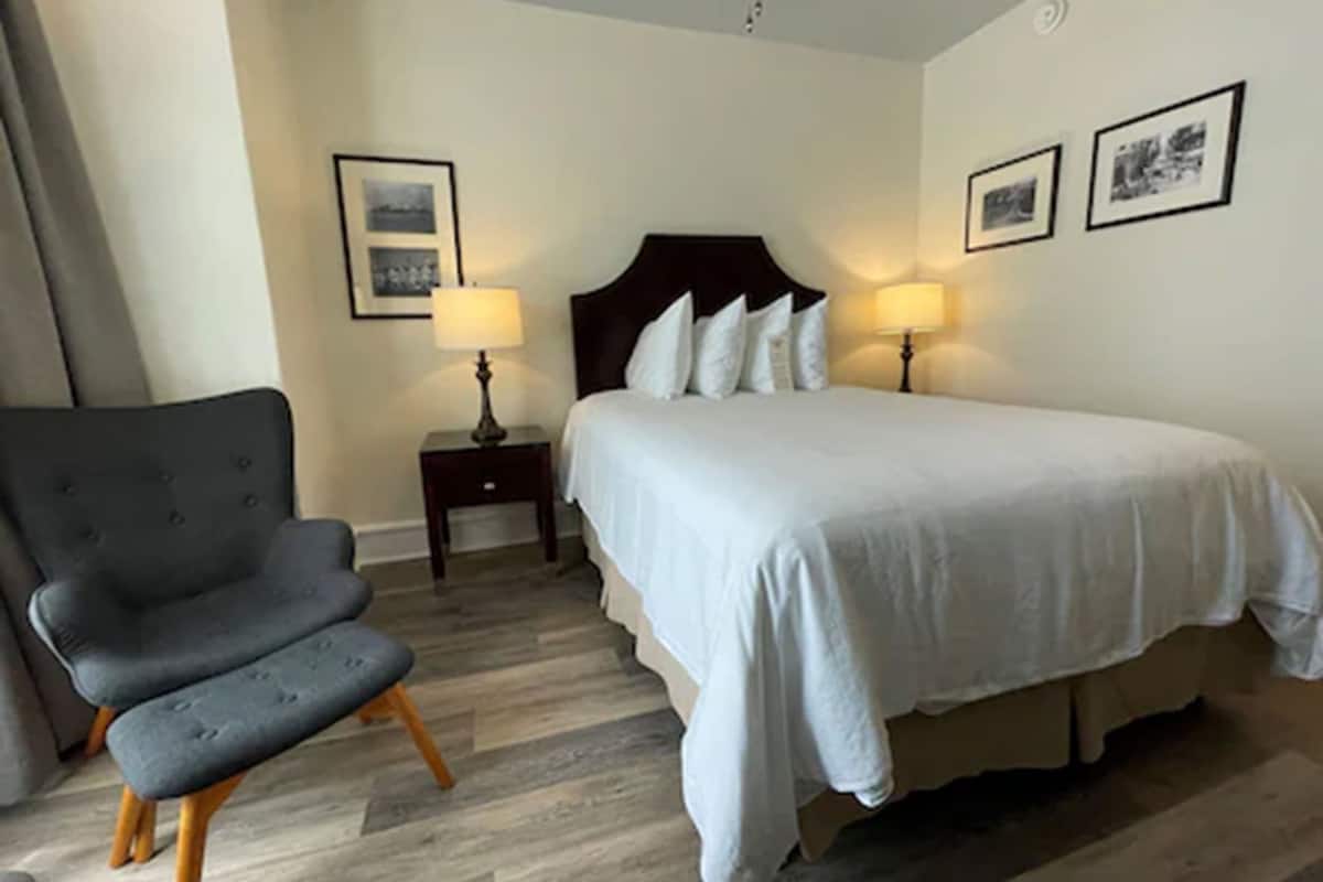 A hotel room with a large bed, two bedside tables with lamps, a grey armchair, and a wooden floor.