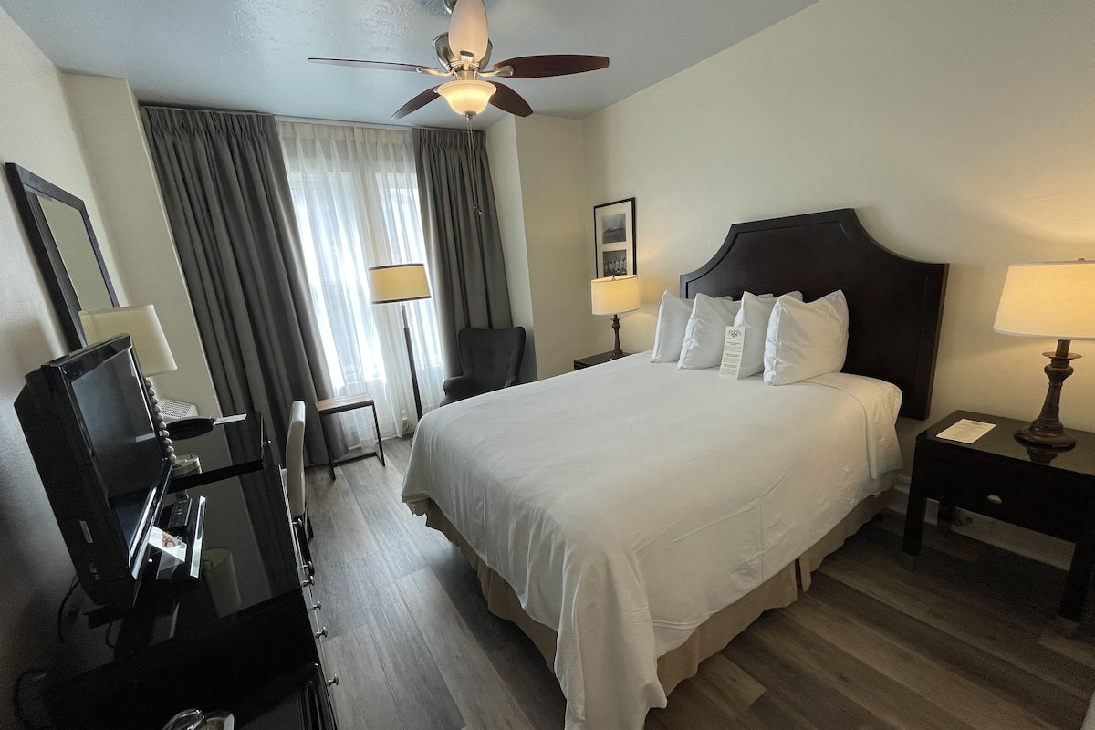 A hotel room with a large bed, a desk with a computer, a chair, a desk lamp, and a ceiling fan.