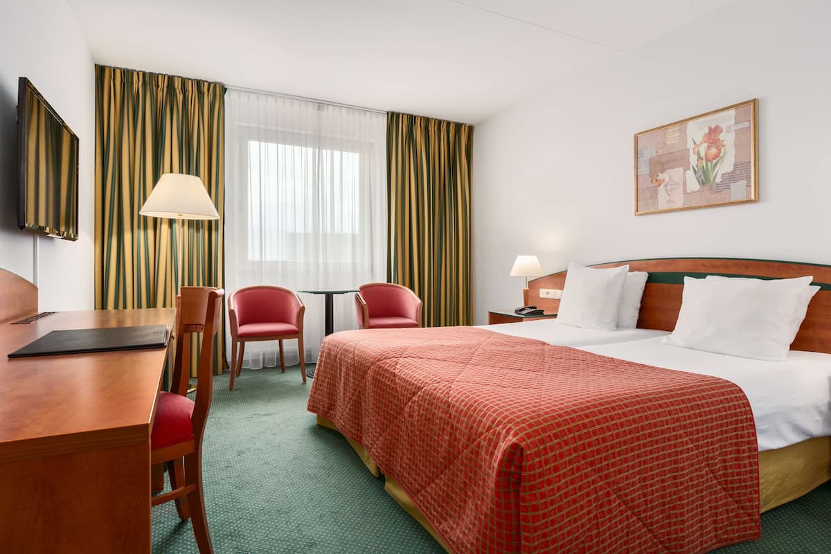 A hotel room with a bed, two chairs, a desk, a lamp, and a painting on the wall.