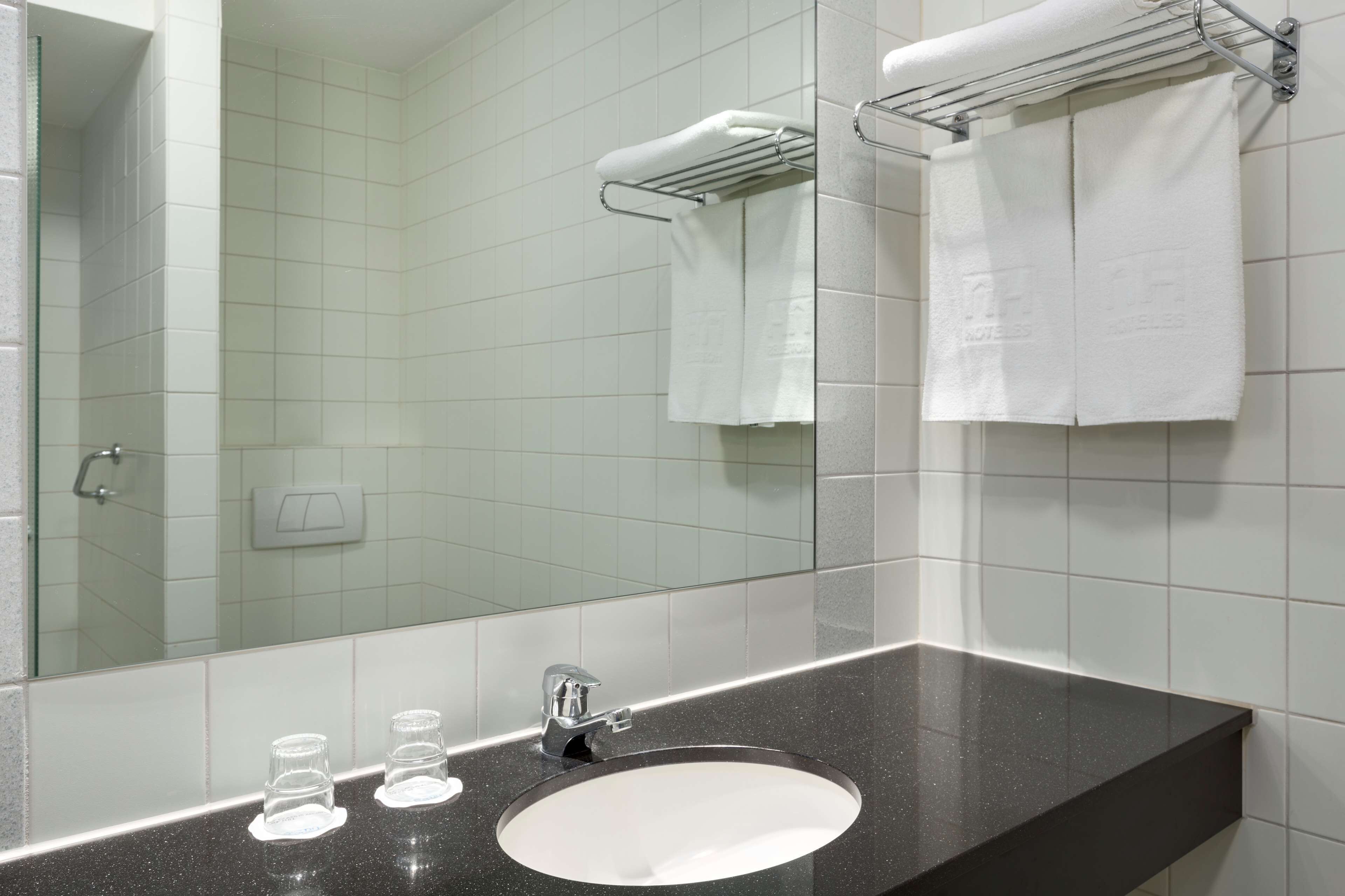 A bathroom with a black countertop, white sink, and a mirror reflecting a towel rack and a shower area.
