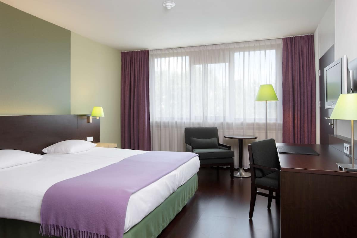 A hotel room with a large bed, two chairs, a desk, a TV, and a window with curtains.