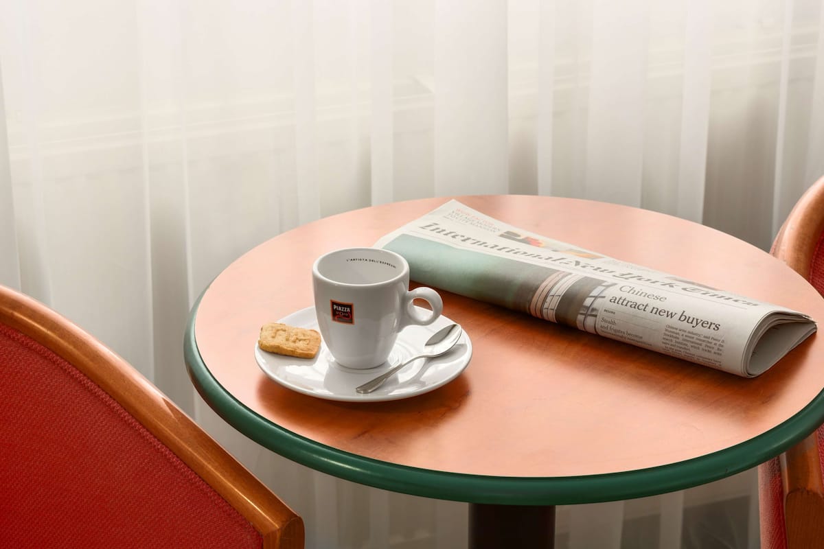 A round glass table with a white mug and a biscuit on a saucer, a newspaper, and a red chair.